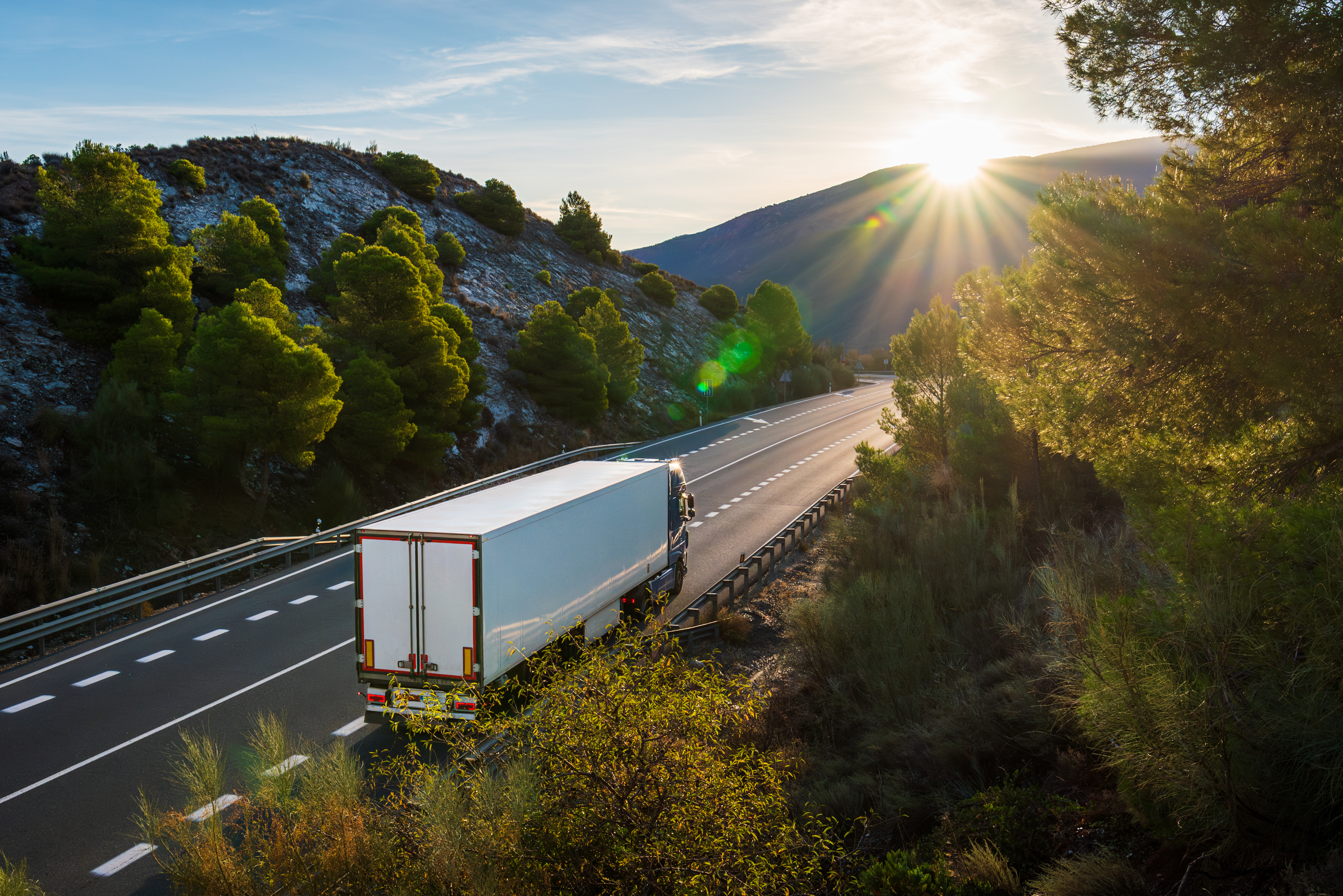 Refrigerated truck driving along a mountain road surrounded by forests, with sunlight breaking over the horizon above the peaks.
